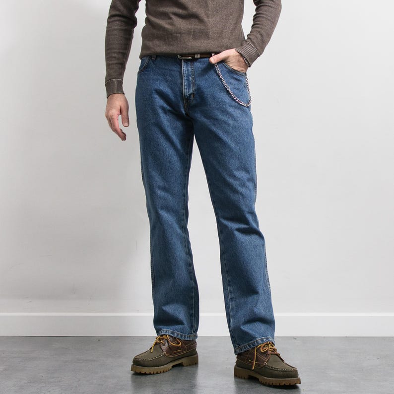 May include: Blue denim jeans with a chain attached to the front pocket. The jeans are paired with a brown long-sleeve shirt and brown and green boat shoes. The outfit is casual and suitable for everyday wear.