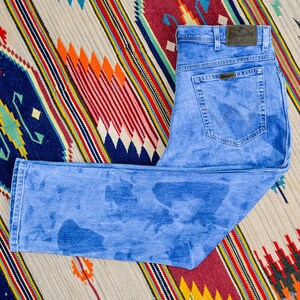 K&ouml;nnte beinhalten: Eine blaue Jeans aus Denim mit verblassem Waschgang und einem Lederpatch mit dem Wrangler-Logo am hinteren Bund.