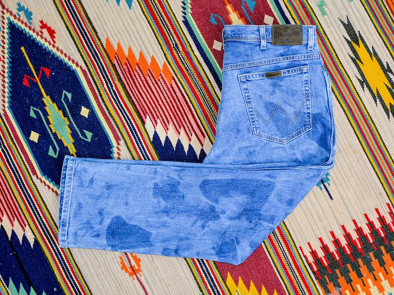 K&ouml;nnte beinhalten: Eine blaue Jeans aus Denim mit verblassem Waschgang und einem Lederpatch mit dem Wrangler-Logo am hinteren Bund.
