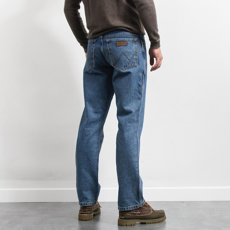 May include: Blue denim jeans with a brown leather patch on the back pocket. The jeans are a classic fit and are paired with brown leather shoes. The person is wearing a brown long-sleeve shirt.