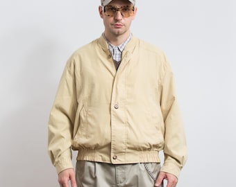 May include: A tan bomber jacket with a button closure, worn over a collared shirt and a sweater. The person is wearing a baseball cap and sunglasses. The jacket has a ribbed hem and cuffs. The pants are khaki-colored.