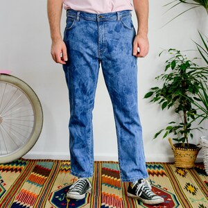 K&ouml;nnte beinhalten: Eine Person tr&auml;gt eine blaue Jeans mit einem verblassten Batik-Muster. Die Jeans wird mit schwarzen Sneakern getragen und die Person steht auf einem bunten Teppich.