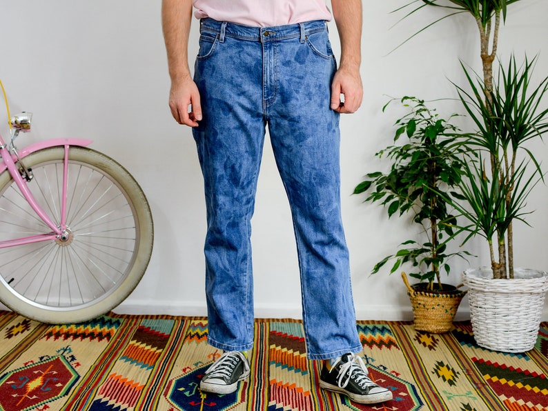 K&ouml;nnte beinhalten: Eine Person tr&auml;gt eine blaue Jeans mit einem verblassten Batik-Muster. Die Jeans wird mit schwarzen Sneakern getragen und die Person steht auf einem bunten Teppich.