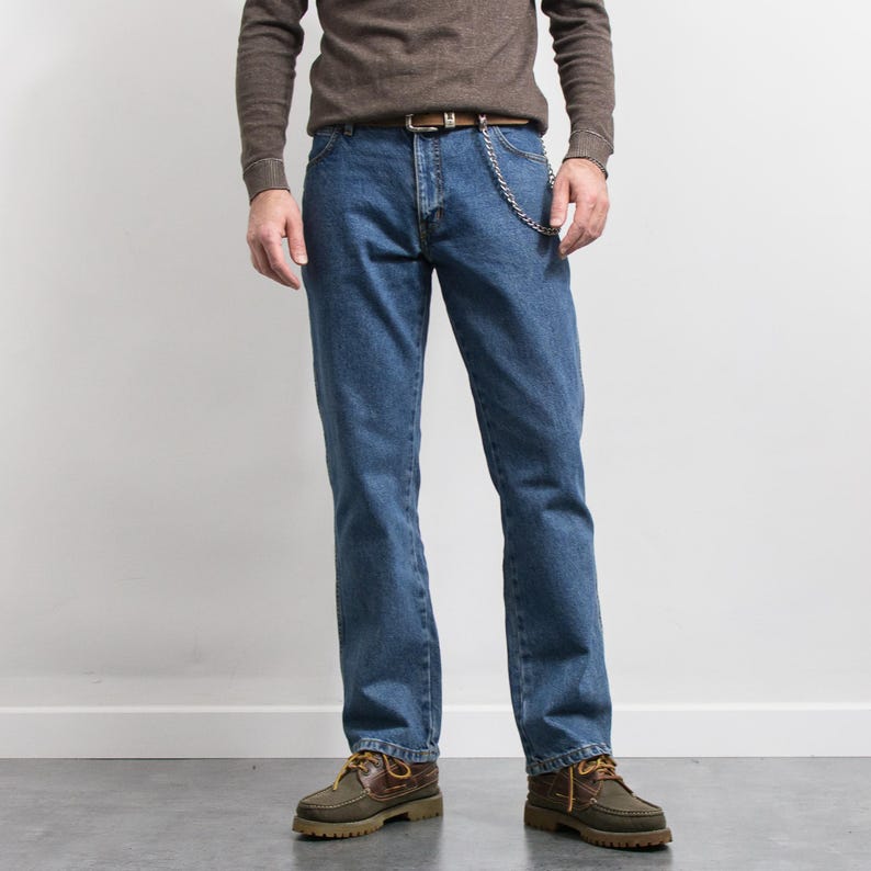 May include: Blue denim jeans with a brown leather belt and a silver chain. The jeans are paired with a brown long-sleeve shirt and brown and green shoes. The jeans have a classic, straight-leg fit.