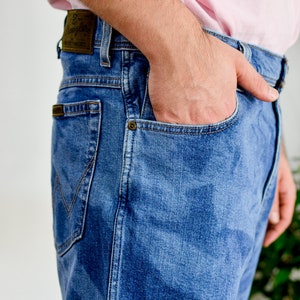 K&ouml;nnte beinhalten: Nahaufnahme einer Person, die eine blaue Jeans aus Denim mit einem braunen Leder-Patch tr&auml;gt, auf dem "Wrangler" auf der Ges&auml;&szlig;tasche steht. Die Hand der Person befindet sich in der vorderen Tasche.