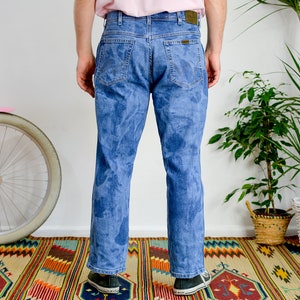 K&ouml;nnte beinhalten: Eine blaue Jeans mit einem Batik-Muster. Die Jeans wird von einer Person getragen, die auf einem bunten Teppich steht.