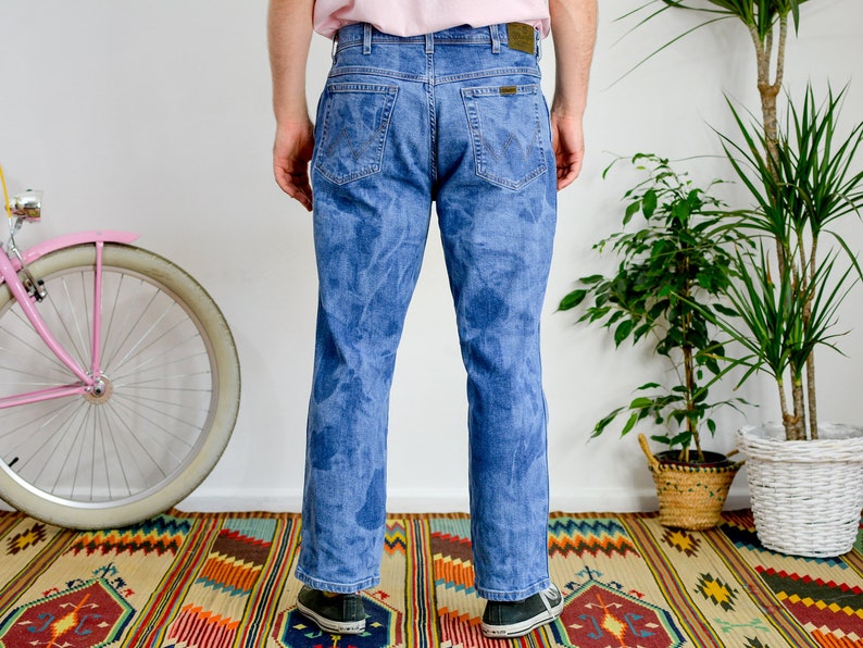 K&ouml;nnte beinhalten: Eine blaue Jeans mit einem Batik-Muster. Die Jeans wird von einer Person getragen, die auf einem bunten Teppich steht.