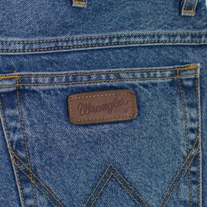 May include: Close-up of the back pocket of blue denim jeans. The jeans have yellow stitching and a brown leather patch with the brand name "Wrangler" embossed on it. The denim has a faded, worn appearance.