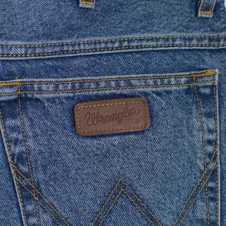 May include: Close-up of the back pocket of blue denim jeans. The jeans have yellow stitching and a brown leather patch with the brand name "Wrangler" embossed on it. The denim has a faded, worn appearance.