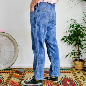 K&ouml;nnte beinhalten: Eine Person tr&auml;gt eine blaue Jeans mit einem verblassten Batik-Muster. Die Jeans ist locker geschnitten und hat ein gerades Bein. Die Person tr&auml;gt schwarze Turnschuhe.