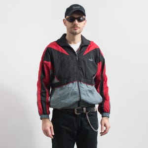 Puede incluir: Chaqueta deportiva Puma vintage en negro, rojo y gris. La chaqueta tiene cremallera, cuello alzado y el logo de Puma en el pecho. La chaqueta se usa con pantalones negros y una gorra negra.
