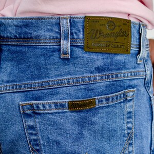 K&ouml;nnte beinhalten: Blaue Jeans aus Denim mit einem braunen Leder-Wrangler-Etikett auf der Ges&auml;&szlig;tasche. Das Etikett tr&auml;gt die Aufschrift "Wrangler Durable Quality".