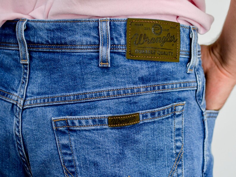 K&ouml;nnte beinhalten: Blaue Jeans aus Denim mit einem braunen Leder-Wrangler-Etikett auf der Ges&auml;&szlig;tasche. Das Etikett tr&auml;gt die Aufschrift "Wrangler Durable Quality".
