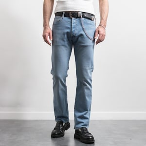 Op de afbeelding: Lichtblauwe spijkerbroek met een klassiek ontwerp met vijf zakken. De jeans wordt gedragen met een zwarte leren riem en een zilveren ketting. Het model draagt ook zwarte leren loafers.