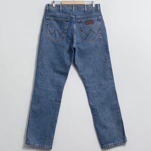 May include: Light blue denim jeans hanging on a wooden hanger. The jeans have a classic five-pocket design with the signature "W" stitching on the back pockets and a brown leather patch. The jeans are a straight-leg style.