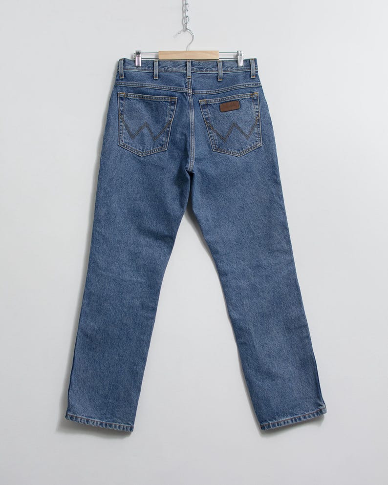 May include: Light blue denim jeans hanging on a wooden hanger. The jeans have a classic five-pocket design with the signature "W" stitching on the back pockets and a brown leather patch. The jeans are a straight-leg style.