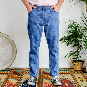 K&ouml;nnte beinhalten: Eine Person tr&auml;gt eine blaue Jeans mit einem verblassten Batik-Muster. Die Jeans ist gerade geschnitten und hat eine lockere Passform. Die Person steht auf einem bunten Teppich mit einem rosafarbenen Fahrrad im Hintergrund.