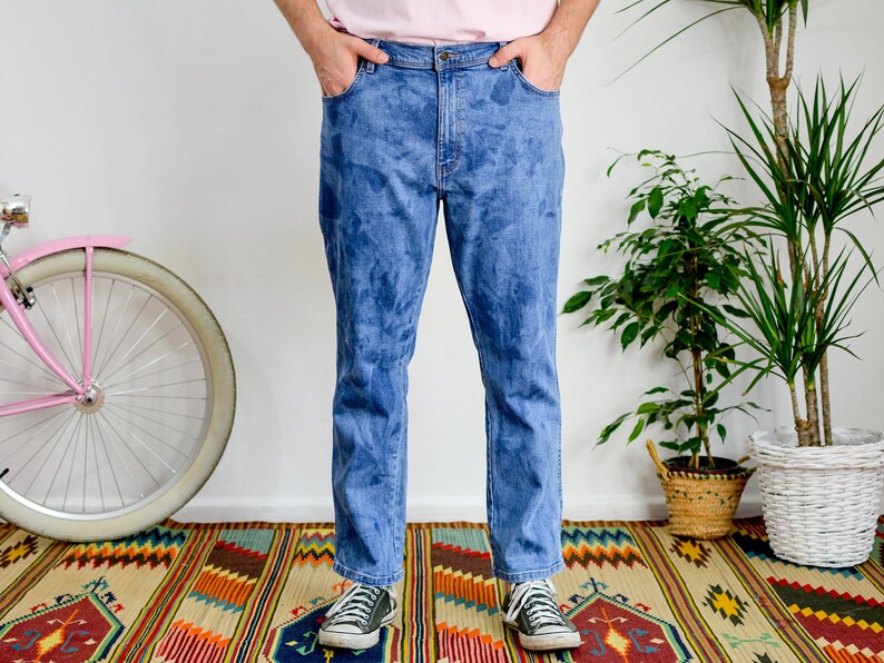 K&ouml;nnte beinhalten: Eine Person tr&auml;gt eine blaue Jeans mit einem verblassten Batik-Muster. Die Jeans ist gerade geschnitten und hat eine lockere Passform. Die Person steht auf einem bunten Teppich mit einem rosafarbenen Fahrrad im Hintergrund.