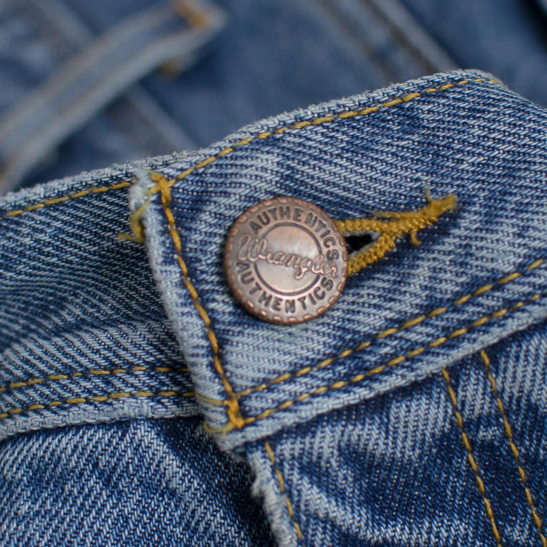 May include: Close-up of blue denim fabric with a copper-colored button. The button is embossed with the words "Wrangler Authentics" in a circular pattern. Yellow stitching accents the denim.