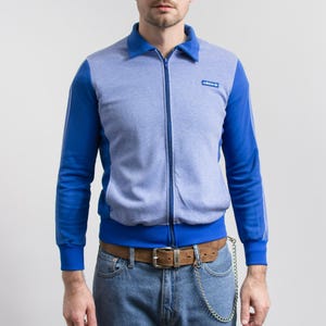 Puede incluir: Chaqueta deportiva azul y gris con cremallera, cuello y logotipo de Adidas. La chaqueta tiene mangas azules con rayas blancas y un dobladillo y puños acanalados azules. El modelo lleva vaqueros azules y un cinturón marrón.