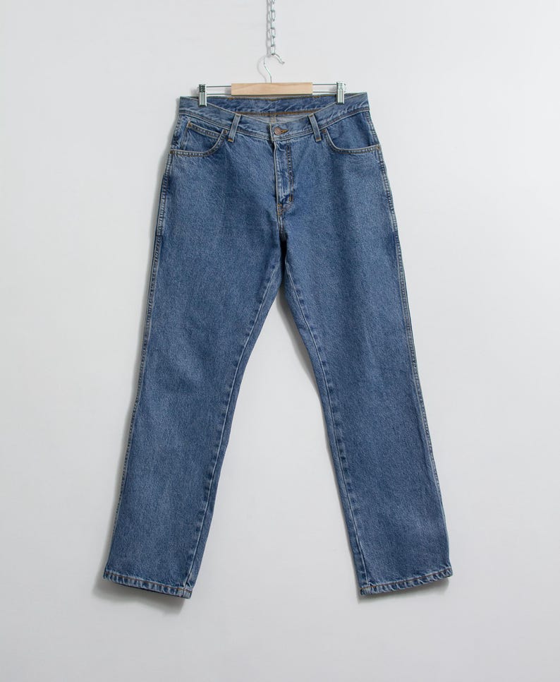 May include: A pair of light blue denim jeans hangs from a wooden hanger. The jeans have a classic five-pocket design, a button and zipper closure, and a straight leg cut. The denim has a medium wash with subtle fading.