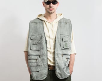 Puede incluir: Un chaleco vaquero verde claro con múltiples bolsillos y cremalleras. El chaleco se lleva sobre una sudadera con capucha color crema. La persona lleva una gorra de béisbol y gafas de sol. El chaleco tiene un estilo casual y utilitario.