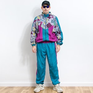 Op de afbeelding: Een vintage teal en paars trainingspak. De jas heeft een kleurrijk patroon met paarse en teal accenten, een teal rits en een paars middenpaneel. De bijpassende broek is teal met paarse strepen aan de zijkanten. De outfit komt uit de jaren 80 of 90.