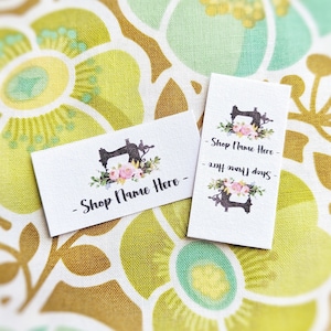 May include: Two white rectangular tags with black text and a black and white sewing machine graphic with pink flowers. The text on the tags reads "Shop Name Here - Shop Here Now". The tags are on a floral fabric background.