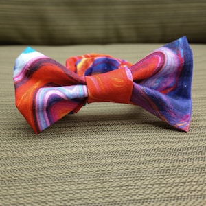 May include: A handmade bow tie with a vibrant, swirling pattern in shades of red, orange, purple, and blue. The bow tie is made of fabric and is designed to be worn around the neck. The bow tie is a stylish accessory.