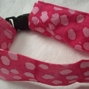 May include: A pink fabric collar with a white spotted pattern and a black plastic buckle. The collar is designed for a pet, likely a dog or cat. The fabric is a vibrant pink.