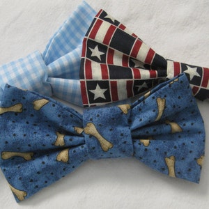 May include: Three fabric bow ties in the image. One is light blue gingham, another has a blue background with tan dog bones, and the third has a patriotic red, white, and blue star and stripe pattern.