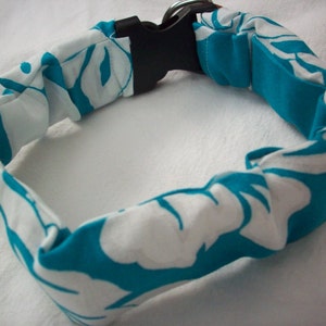 May include: A fabric dog collar with a white and teal floral pattern. The collar has a black plastic buckle and a metal D-ring. The collar is made of a soft, flexible material.