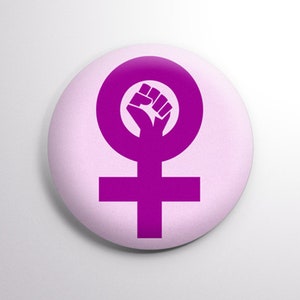 May include: A purple button with a raised image of a female symbol with a fist inside the circle. The background of the button is a light pink.