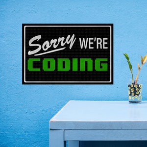 May include: Black and white sign with green text that reads "Sorry We're Coding" on a blue wall. The sign is surrounded by a pattern of 0s and 1s.