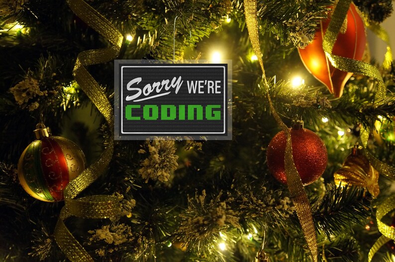 May include: A Christmas tree decorated with ornaments and gold ribbon. A rectangular sign with the words "Sorry We're Coding" in white and green is hanging on the tree.