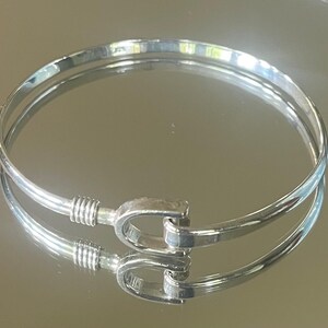 May include: A silver-colored bangle bracelet with a hook clasp. The bracelet is round and has a smooth, polished finish. The clasp is a U-shaped design. The bracelet is displayed on a reflective surface.