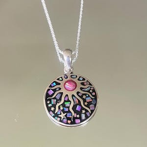 May include: A circular silver pendant on a silver chain featuring a central pink cabochon surrounded by a sun motif. The background of the pendant is black and inlaid with small, iridescent, multi-colored mosaic pieces and tiny silver stars.