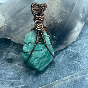 May include: A wire-wrapped pendant featuring a rough-cut, light blue-green gemstone. The pendant is encased in copper-colored wire, with intricate wrapping details at the top and around the stone. The gemstone has a textured surface.