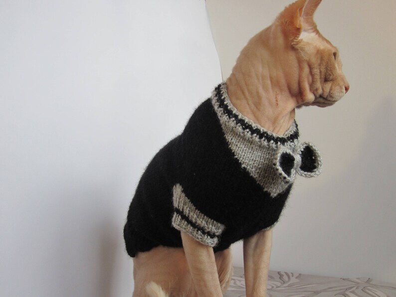 Pu&ograve; includere: Un gatto senza pelo che indossa un maglione di lana nero e grigio con un papillon. Il maglione ha un colletto e polsini grigi.