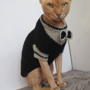 Pu&ograve; includere: Un gatto senza pelo che indossa un maglione di lana nero e grigio con un papillon. Il gatto &egrave; seduto su una superficie con un motivo.