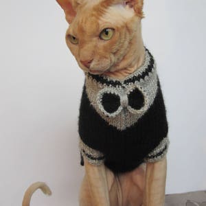 Pu&ograve; includere: Un gatto senza pelo che indossa un maglione di lana nero e grigio con un papillon. Il gatto &egrave; seduto su una superficie con un motivo.