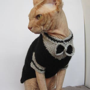 Pu&ograve; includere: Un gatto senza pelo che indossa un maglione di lana nero e grigio con un papillon. Il gatto &egrave; seduto su una superficie a motivi.
