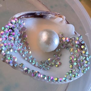 Puede incluir: Una concha blanca con una perla en el centro y piedras de imitación iridiscentes alrededor del borde.