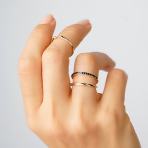 Può includere: Tre anelli d'oro su una mano. L'anello superiore è una semplice fascia curva. L'anello centrale è una fascia sottile e liscia. L'anello inferiore è una fascia sottile con pietre nere.