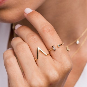 Può includere: Una mano di donna con due anelli d'oro. Uno è una semplice fascia con una piccola pietra chiara. L'altro anello è una forma geometrica con una punta.