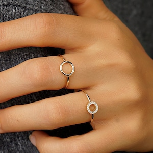 Könnte beinhalten: Zwei goldene Ringe an einer Hand. Ein Ring ist ein einfacher Kreis, der andere ist ein Kreis mit kleinen Diamanten am Rand.