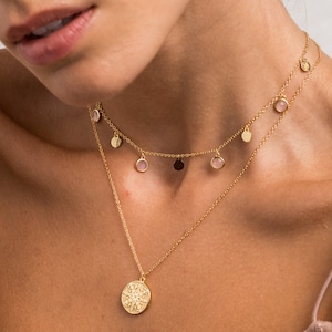 以下が含まれることがあります： 繊細なチェーンと、詳細なデザインが施された丸いペンダントが付いたゴールドネックレス。ネックレスには、小さな丸いピンクの石がぶら下がった2番目のチェーンも付いています。