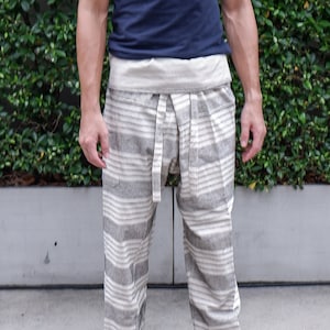 May include: A person wearing a navy blue crew neck t-shirt and striped gray and white pants with a drawstring waist. The pants have a relaxed fit and are made of a lightweight fabric. The background is a green hedge.