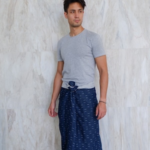 以下が含まれることがあります： グレーのTシャツと、白い水玉模様のネイビーラップスカートを着用した男性。スカートは、お揃いの生地でウエストに結ばれています。彼はまた、黒いビーチサンダルを履いています。