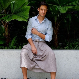 以下が含まれることがあります： ライトブルーのボタンダウンシャツとトープ色のパンツを着た人が、コンクリートの縁に座っています。その人は茶色のスエードの靴を履いています。背景には緑豊かな葉があります。